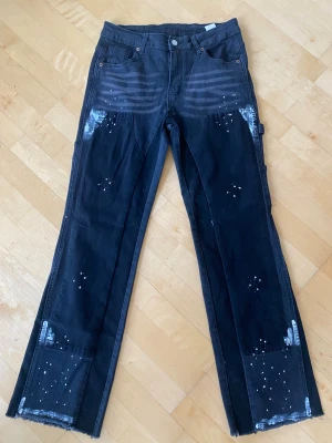 Svarta straight leg jeans med färgstänk - Säljer ett par svarta straight leg jeans med råa benslut och vita färgstänk för en cool, avslappnad look. Jeansen har flera fickor, inklusive sidofickor och bakfickor, samt detaljer som slitningar och penselfäste. Perfekta för dig som gillar streetwear och unik stil.