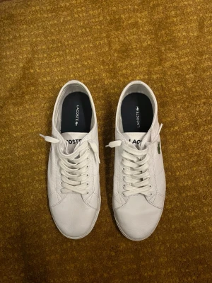 Vita Lacoste sneakers i läder - Säljer ett par klassiska vita sneakers från Lacoste i läder med snörning och den ikoniska gröna krokodilloggan på sidan. Skorna har låg profil och stilren design som passar till det mesta. Perfekta för dig som gillar clean och tidlös stil.