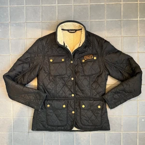 Fodrad vårjacka av barbour - Snygg svart quiltad jacka från Barbour med guldfärgade knappar och fyra fickor framtill. Jackan har en mjuk, vit teddyfodrad krage och detaljerad Barbour-logga på bröstet. Perfekt för dig som vill ha en klassisk men trendig look.