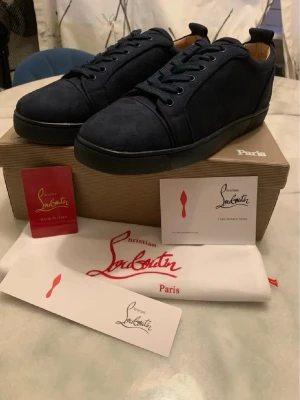 Christian Louboutin skor  - Snygga mörkblå sneakers från Christian Louboutin i mocka med klassisk röd sula. Skorna har rund tå, matchande mörkblå skosnören och diskreta sömmar. Insidan är fodrad i ljust läder och designen är clean och stilren. Perfekta för dig som gillar exklusiva detaljer.
