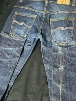 Blå jeans från Nudie Jeans - Säljer ett par klassiska blå jeans från Nudie Jeans med snygga slitningar och kontrastsömmar. Jeansen har raka ben, två bakfickor med Nudie Jeans signaturbrodyr och en patch i läder bak i midjan. Perfekta för en avslappnad och trendig look.