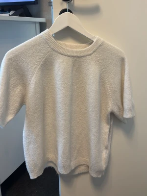 Vit kortärmad stickad tröja - Mysig vit stickad tröja med korta ärmar och rund halsringning. Tröjan har en avslappnad passform och ribbade kanter vid hals, ärmslut och nederkant. Perfekt för lager på lager och enkel att matcha med olika stilar.