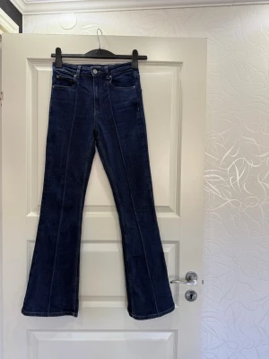 Mörkblå bootcut jeans med söm - Snygga mörkblå bootcut jeans med markerad mittsöm framtill. Klassisk femficksmodell och hög midja. Jeansen har en knapp och dragkedja i gylfen samt kontrastsömmar som ger en trendig look. Perfekta för dig som gillar retrovibbar och vill ha ett par jeans som sticker ut.