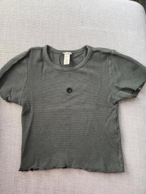 Ribbad grön croppad topp från Pull&Bear - En mörkgrön croppad topp från Pull&Bear med ribbad struktur och korta ärmar. Toppen har en liten broderad svart blomma på bröstet. Hel och ren och i mycket bra skick. Storlek: S