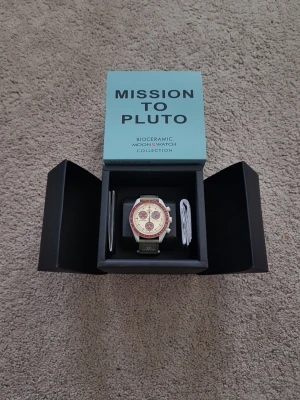 Swatch x Omega Mission to Pluto klocka - Säljer en Swatch x Omega Mission to Pluto från Bioceramic MoonSwatch-kollektionen. Klockan har en vit boett, vinröd bezel och detaljer, samt grått armband. Snygg och unik design med kronograf och tydliga knappar på sidan. Kommer i originalbox.