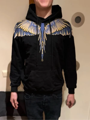 Marcelo Burlon svart hoodie med vingar - Svart hoodie från Marcelo Burlon County of Milan med ikoniskt vingtryck i blått och gult över axlar och rygg. Tröjan har huva med snörning och känguruficka framtill. Perfekt statement-plagg för dig som vill sticka ut.