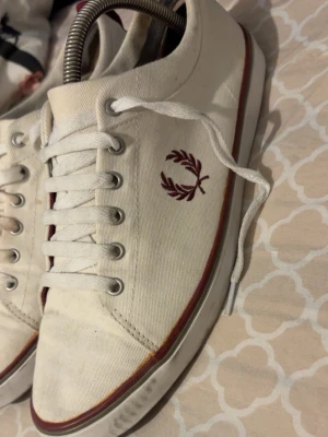 Vita Fred Perry canvas sneakers - Snygga vita sneakers från Fred Perry i canvas med klassisk vinröd logga på sidan och matchande rand längs sulan. Lågt skaft, vita skosnören och metallöljetter ger en clean och stilren look. Perfekta för dig som gillar enkel och tidlös stil.