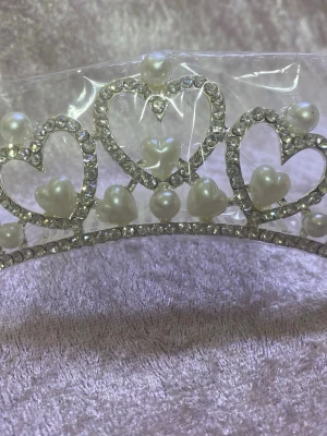 Glittrigt diadem med pärlor och hjärtan - Säljer ett diadem i silverfärg med gnistrande strass och vita pärlor. Diademet har hjärtformade detaljer och pärlor i olika storlekar, vilket ger en romantisk och lyxig känsla. Perfekt för dig som vill sticka ut med en elegant accessoar. 168kr/st 