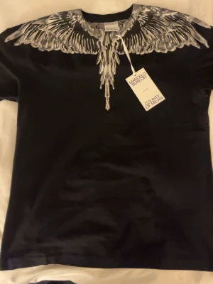  Marcelo Burlon  - Cool svart t-shirt från Marcelo Burlon med ett unikt vitt vingmönster runt halsen. T-shirten har korta ärmar och är tillverkad i mjuk bomull. Perfekt för dig som vill sticka ut med en stilren och edgy look. 