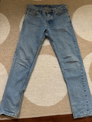 Ljusblå raka jeans från Nudie - Säljer ett par klassiska ljusblå jeans från Nudie med rak passform och fem fickor. Jeansen har en tidlös look med subtila detaljer på bakfickorna och är tillverkade i 100% bomull. Perfekta för en avslappnad och trendig stil. W28 L30