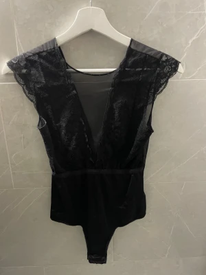 Svart spetsbody med djup urringning - Snygg svart body med spetsdetaljer och djup v-ringning framtill. Axelpartiet har transparent mesh och spets.
