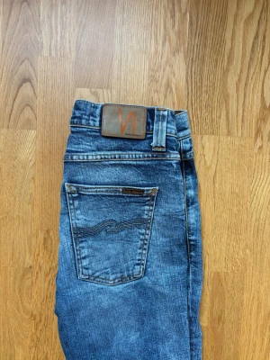 Blå jeans från Nudie Jeans - Säljer ett par klassiska blå jeans från Nudie Jeans med snygg tvätt och smal passform. Jeansen har fem fickor, läderpatch med logga bak och coola detaljer på bakfickan. Perfekta för dig som gillar en avslappnad men stilren look.
