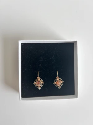 Guldtonade örhängen med pärldetaljer - Original pris 1000 kr. Använt en gång. Unika örhängen i guldton med rund form och dekorativa pärldetaljer. De har en vintageinspirerad design med små vita pärlor och en central sten i varm rosa nyans. Perfekta för dig som gillar detaljerade accessoarer med retrovibbar.