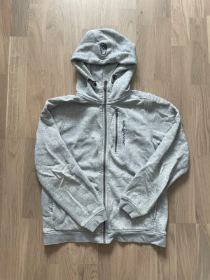 Grå zip-hoodie från Sail Racing - Snygg grå hoodie från Sail Racing med dragkedja framtill och huva med dragsko. Märkets logga syns på bröstet och huvan. Hoodien har ribbade muddar vid ärmslut och nederkant samt en bröstficka med dragkedja. Perfekt för en avslappnad streetwear-look.