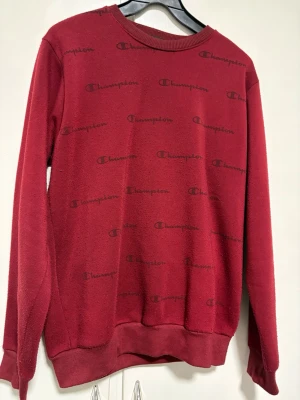 Röd sweatshirt från Champion - Säljer en röd sweatshirt från Champion med rund halsringning och svarta logotyper tryckta över hela tröjan. Tröjan har långa ärmar, ribbade muddar och är gjord i mjukt sweatshirtmaterial. Perfekt för en avslappnad och sportig stil.