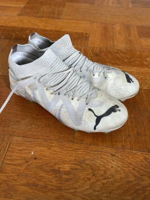Vita Puma fotbollsskor med högt skaft - Säljer ett par vita fotbollsskor från Puma med högt stickat skaft och snörning. Skorna har svart Puma-logga på ovansidan och mönstrad sula med gröna dobbar. Ovandelen är i syntetmaterial och designen är modern och sportig.