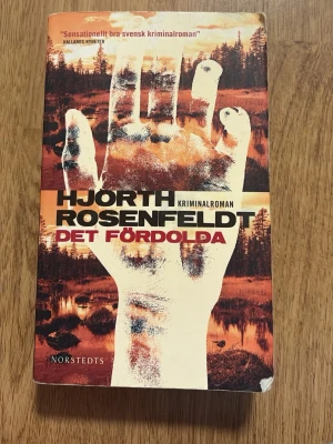 Det fördolda av Hjorth och Rosenfeldt - Spännande kriminalroman av Hjorth, Rosenfeldt. Omslaget har en stor vit hand mot en mörk skog och orange bakgrund. Boken är i pocketformat och har tydliga svarta och röda titlar på framsidan. Perfekt för dig som gillar svenska deckare. Första boken i serien!