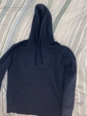 Mörkblå hoodie från Boss - Säljer en mörkblå hoodie från Boss med broderad logga på bröstet. Hoodien har en stor huva med dragsko och en klassisk magficka framtill. Tillverkad i mjukt material som känns skönt mot huden. Perfekt för en avslappnad och stilren look.