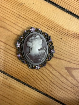 Kamé medaljong - Hänge till halsband i lila och vitt samt mörk metall med små glittriga stenar (några har lossnat).  Hängögla sitter på baksidan.