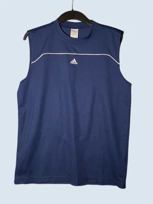 Blå ärmlös linnetopp från Adidas - Sportig blå linnetopp från Adidas med vitt diskret logotryck på bröstet och vit linje över bröstet. Tillverkad i lätt och funktionell polyester, perfekt för träning eller en aktiv dag. Klassisk rund halsringning och avslappnad passform.