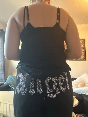 Svarta mjukisbyxor med strassdetalj - Säljer ett par svarta mjukisbyxor med resår i midjan och dragsko. På baksidan står det 'Angel' i stora bokstäver av strass, vilket ger en snygg och trendig look. Perfekta för dig som gillar statement-plagg och vill ha något bekvämt men ändå stilrent.