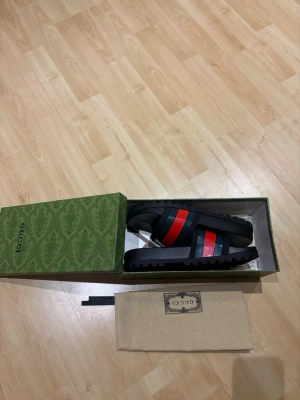Svarta sandaler från Gucci - Säljer ett par svarta sandaler från Gucci med klassiska gröna och röda ränder över foten. Sandalerna har en bred rem i gummi och en räfflad sula för extra grepp. Kommer i originalkartong och med dustbag.