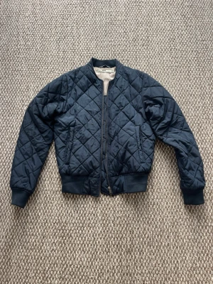 Marinblå quiltad bomberjacka HOPE - Marinblå quiltad bomberjacka från HOPE med ribbade muddar och dragkedja framtill. Jackan har ett klassiskt rutmönster och rundad krage. 