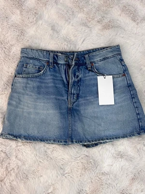 Blå jeanskjol från Zara  - HELT NY MED PRISLAPP Trendig blå jeanskjol i kort modell med rå kant nertill. Kjolen har klassiska fem fickor, bälteshällor och knapp- samt dragkedjestängning framtill. Perfekt för en avslappnad och cool stil.