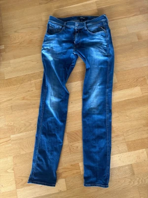 Blå slim fit Anbass  jeans från Replay (w32/l34) - Säljer ett par blå slim fit Anbass jeans från Replay (w32/l34) med snygga slitningar och klassiska fem fickor. Jeansen har en skinnpatch bak i midjan och kontrastsömmar. Perfekta för en avslappnad och trendig look.