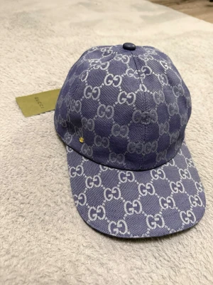 Blå Gucci keps med GG-mönster - Säljer en blå keps från Gucci med det klassiska GG-monogrammet i vitt över hela kepsen. Kepsen har böjd skärm och justerbart band bak i blått läder. Materialet är canvas och kepsen har en exklusiv känsla med märkets tag kvar.