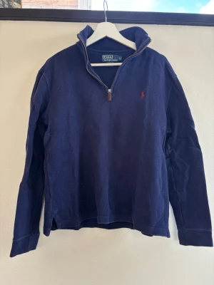 Marinblå halvzip från Ralph Lauren - Klassisk marinblå tröja från Polo Ralph Lauren med halvzip och hög krage. Tröjan har lång ärm och den ikoniska röda loggan broderad på bröstet. Tillverkad i mjukt bomullsmaterial, perfekt för en avslappnad men stilren look.