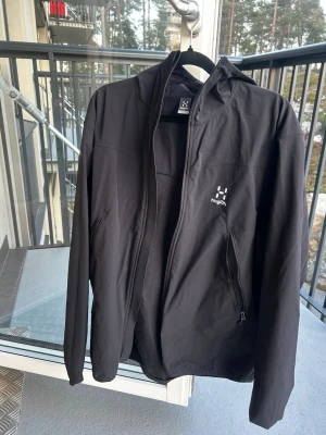 Svart vindjacka från Haglöfs - Snygg svart vindjacka från Haglöfs med dragkedja framtill och två sidofickor med dragkedja. Jackan har en minimalistisk design, luva och diskret logga på bröstet. Perfekt för blåsiga dagar och enkel att matcha med olika outfits.