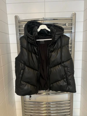 Svart dunväst med huva från Zara - Snygg svart dunväst från Zara med dragkedja, stora fickor och en rejäl huva med dragsko. Västen har ett quiltat mönster.Längst ner på västen finns en dragsko(se bild) som man kan justera. Perfekt att slänga över en hoodie för extra värme.