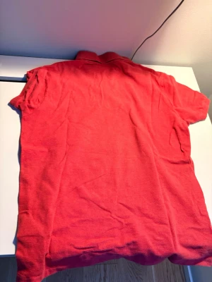 Röd polotopp från Polo Ralph Lauren - Säljer en klassisk röd polotopp från Polo Ralph Lauren med korta ärmar och krage. Toppen har knappar framtill och den ikoniska logon broderad på bröstet. Perfekt för dig som gillar en sportig och stilren look.