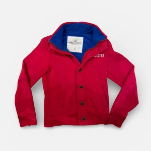 Hollister vintage (Y2K) - Säljer en riktigt snygg och eftertraktad Hollister-tröja i en klar röd färg med marinblått foder i kragen. Det här är en klassisk vintage-modell som inte längre går att köpa i butik! Tröjan har både dragkedja och snygga knappar framtill