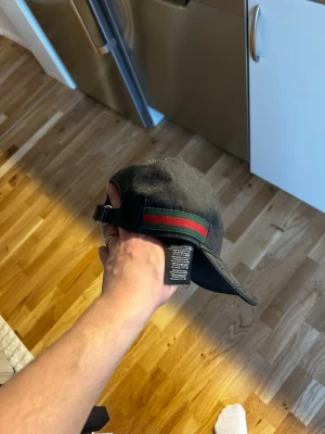 Svart Gucci keps med röd-gröna ränder - Säljer en svart keps från Gucci med klassiska röd-gröna ränder på sidan. Kepsen har justerbart spänne bak och diskret GG-mönster över hela tyget. Perfekt för dig som vill ha en stilren och lyxig accessoar till din outfit.