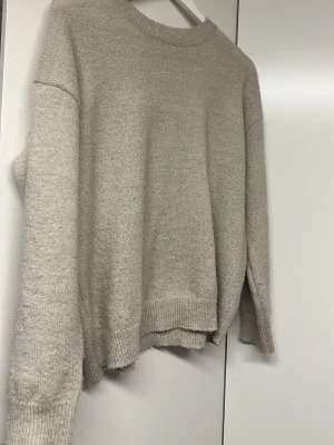 Beige stickad tröja i enkel design - Mysig beige stickad tröja med rund hals och långa ärmar. Tröjan har en avslappnad passform och ribbade muddar vid ärmslut och nederkant. Perfekt för lager på lager under kyliga dagar.