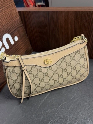 Beige GG canvas axelremsväska  - Snygg beige axelremsväska  i klassiskt GG canvasmönster med detaljer i ljust läder. Väskan har guldfärgade metalldetaljer, kedjerem och dragkedja upptill. Perfekt storlek för det viktigaste och en ikonisk accessoar för dig som gillar lyxig stil.