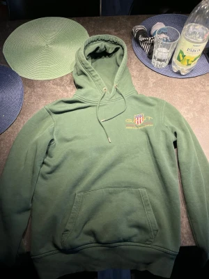 Grön hoodie från GANT - Mysig grön hoodie från GANT med broderad logga och texten 'American Sportswear' på bröstet. Klassisk känguruficka framtill, justerbar huva med dragsko och ribbade muddar. Perfekt för en avslappnad och sportig stil.