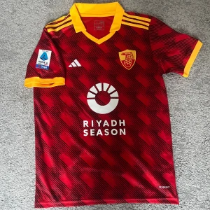 Röd fotbollströja från Adidas - Stilig AS Roma tröja som användes i Derbyt mot Lazio förra säsongen. Tröjan har det gamla emblemet som ger det en liten mer klassisk stil på det hela. Itialenaren Lorenzo Pellegrini är på ryggen och Serie A trycket på tröjärmen finns även med. Tröjan är knappt använd och är alltså i helt perfekt skick. Den är i storlek S, men kan även passa om man har M.