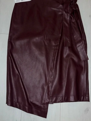 Vinröd omlottkjol i skinnimitation - Snygg vinröd omlottkjol i skinnimitation med knytband i midjan. Kjolen har en lätt glansig finish och en stilren, rak passform som ger en trendig look. Perfekt att matcha med en enkel topp eller stickad tröja.