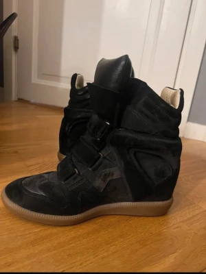 Svarta Isabel Marant wedge sneakers - Säljer ett par svarta wedge sneakers från Isabel Marant med cool design och högt skaft. Skorna har kardborreband, detaljer i mocka och en beige innersida. Sulan är i gummi och ger en snygg kontrast till det svarta. Perfekta för dig som vill ha en edgy och trendig look.
