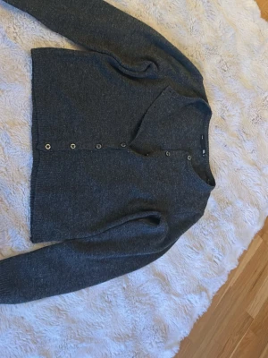 Mörkgrå kofta från 157 - Mysig mörkgrå kofta från 157 med knappar framtill och ribbade muddar. Klassisk rak modell med v-ringning, perfekt att slänga på sig över en t-shirt. Materialet känns mjukt och passar bra till lager-på-lager-stil.