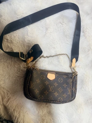 Elegant väska - Snygg handväska från i klassiskt brunt monogrammönster. Väskan har två fack med guldfärgade dragkedjor, en bred svart axelrem och en dekorativ guldkedja. Stilren design som passar perfekt för resor eller vardagsbruk. Den har spår av användning på insidan, vänligen titta noga på bilderna. 