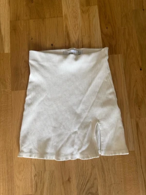 Vit ribbad kortkjol från Bershka - Säljer en vit ribbad kortkjol från Bershka i storlek S. Kjolen har en enkel och clean look med en liten slits framtill som ger en snygg detalj. Perfekt att matcha med crop tops eller oversized tröjor för en trendig stil.