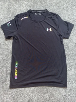 Svart tränings-tshirt från Under Armour - Snygg svart tränings-tshirt från Under Armour med regnbågsfärgad logga på bröstet och texten UNDER ARMOUR i färg längs sidan. Tillverkad i ett lätt och snabbtorkande funktionsmaterial, perfekt för träning och sport.