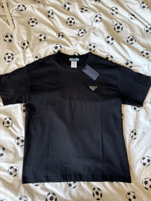 Svart t-shirt från Prada - Snygg svart t-shirt från Prada med klassisk passform och korta ärmar. T-shirten har en diskret logga på bröstet och är tillverkad i mjuk bomull för maximal komfort. Perfekt basplagg med exklusiv känsla.