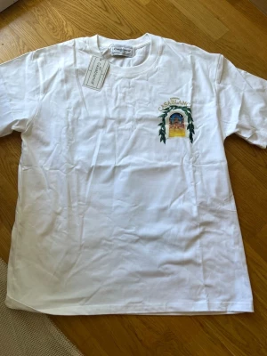 Vit Casablanca t-shirt med tryck - Säljer en vit t-shirt från Casablanca med färgglatt tryck av en byggnad och gröna blad både fram och bak. T-shirten har rund hals och korta ärmar. Perfekt för dig som gillar stilrena plagg med unika detaljer.
