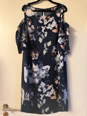 Blommig marinblå offshoulder klänning - Snygg marinblå klänning med blommigt mönster i vitt, rosa och ljusgrönt. Offshoulder-modell med korta, vida ärmar och rak passform. 