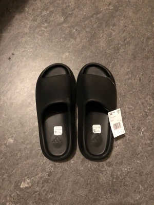 Yeezy Slides - Säljer ett par svarta slides från Adidas med öppen tå och bred rem över foten. Tillverkade i ett mjukt och flexibelt syntetmaterial som ger skön komfort. Perfekta att glida runt i hemma eller på stranden. Klassisk Adidas-logga på sulan.